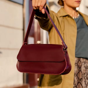Peut inclure: Un sac à bandoulière bordeaux profond avec une finition lisse, un rabat rectangulaire et une anse fixe assortie. Le sac présente des lignes épurées et structurées, mettant en valeur une couleur rouge foncé riche sur tout l'extérieur.