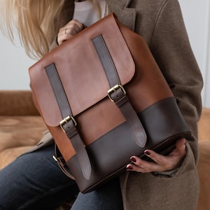 May include: A brown leather backpack with a flap closure and adjustable straps. The backpack has a two-tone design with a darker brown base and accents, and gold-coloured buckles. The bag is held by a person.