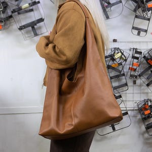 以下が含まれることがあります： 茶色の大きなレザートートバッグ。肩掛けストラップ付き。肩にかけています。茶色のジャケットとサングラスをかけています。背景には白い壁に取り付けられた金属製の構造があります。