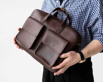 May include: A brown leather briefcase with two front pockets and a top handle. The bag is held by a person wearing a black cap and a blue and white checkered shirt. The bag appears to be new.