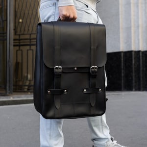 Könnte beinhalten: Ein schwarzer Lederrucksack mit rechteckiger Form und einem Klappverschluss, der mit zwei Riemen und silbernen Schnallen gesichert ist. Der Rucksack wird von einer Person getragen, die helle Jeans und graue Turnschuhe trägt.