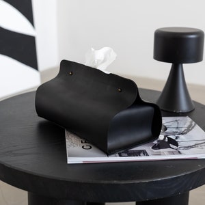 May include: A black leather tissue box cover with a curved design, holding white tissues. It sits on a round, black wooden table with three cylindrical legs, next to a small black decorative object. A magazine is beneath the tissue box.
