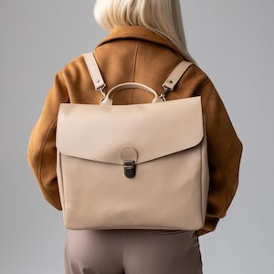 May include: A beige leather backpack with a flap closure and a top handle. The backpack has adjustable straps and a metal clasp. The person is wearing a brown jacket and tan trousers.