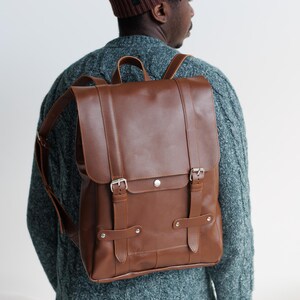 Può includere: Uno zaino in pelle marrone con patta, due cinghie verticali con fibbie in metallo, una maniglia superiore e spallacci regolabili. La borsa presenta una finitura liscia con cuciture visibili e dettagli in metallo color argento.
