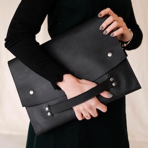 Könnte beinhalten: Schwarze Ledermappe mit Klappe und Griff, befestigt mit silberfarbenen Nieten. Das Design ist schlicht und modern. Das Bild zeigt die Mappe.