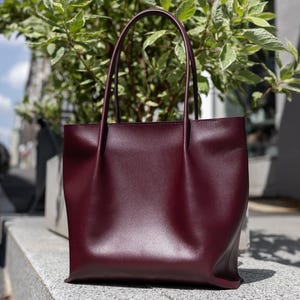 Peut inclure: Un sac cabas bordeaux profond avec une finition lisse et mate, doté de deux anses hautes et fines, ainsi qu'un corps rectangulaire structuré avec de légers plis verticaux sur le panneau avant, créant une silhouette épurée et simple.