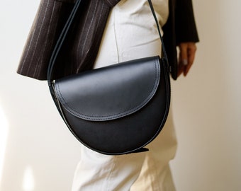 Könnte beinhalten: Eine schwarze Ledersatteltasche mit geschwungenem Design und langem Riemen. Die Tasche wird über der Schulter getragen. Die Person trägt einen braunen Nadelstreifenblazer, ein beiges Oberteil und eine weiße Hose.