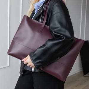 Peut inclure: Un grand sac cabas bordeaux profond avec une finition lisse et mate et deux bandoulières. Le sac présente une silhouette rectangulaire épurée avec une couture horizontale sur la partie inférieure, offrant un design structuré et spacieux.