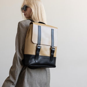 May include: A tan and black leather backpack with two adjustable buckles on the front. The backpack is worn by a person with blonde hair and sunglasses.