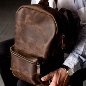 May include: A brown leather backpack with a front zippered pocket and top handle. The backpack has a rounded top and adjustable shoulder straps. The leather has a distressed look. The zipper pulls are leather.