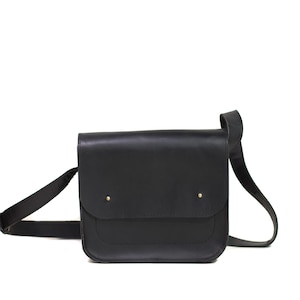 Może przedstawiać: Czarna skórzana torba kurierska z klapką i regulowanym paskiem na ramię. Torba ma dwa małe, złote nity z przodu. Torba jest prostokątna i wydaje się pusta.
