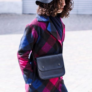May include: A black leather crossbody bag with a flap closure is worn over a plaid coat. The coat features a vibrant pattern of pink, blue, and black. A gray hat is also visible.