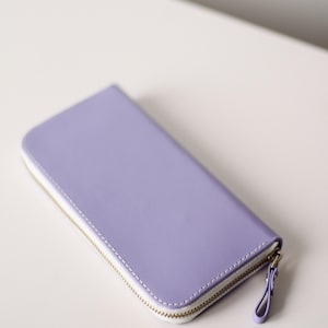 Può includere: Un portafoglio color lavanda con cerniera bianca e finiture color oro. Il portafoglio è rettangolare e sembra essere realizzato in pelle o materiale simile. La cerniera corre su tre lati del portafoglio.