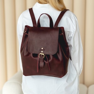 Puede incluir: Mochila de cuero burdeos con cierre de solapa y cordón. La mochila tiene correas ajustables para los hombros, un asa superior y una hebilla dorada. La bolsa se lleva sobre una camisa blanca.