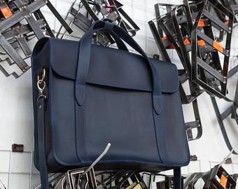 May include: A navy blue leather satchel with a top handle and a detachable shoulder strap. The bag has a flap closure and gold-colored hardware. The background features metalwork hanging on a white grid.