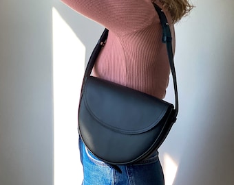 Könnte beinhalten: Eine schwarze Ledersatteltasche mit gebogenem Überschlag und verstellbarem Riemen. Die Tasche wird über der Schulter getragen. Die Person auf dem Bild trägt ein rosa Langarmshirt und blaue Jeans.