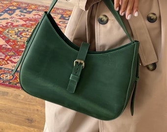 May include: A dark green leather shoulder bag with a curved design. The bag features a strap, a buckle closure, and a gold-colored buckle. The bag is held against a beige trench coat.