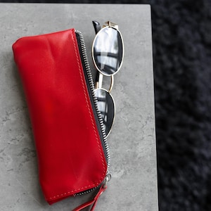 May include: A red leather sunglasses case with a black zipper and a small loop. Gold-rimmed sunglasses with black lenses are next to the case. The case and sunglasses are on a gray surface.
