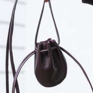 Op de afbeelding: Een kleine, donkerpaarse buidel met trekkoord en een gladde afwerking. De tas heeft een ronde, bolle vorm en een lange, dunne bijpassende schouderband. De opening aan de bovenkant is geplooid en wordt vastgezet met een dun koord.