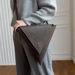 Könnte beinhalten: Eine dunkelbraune Leder-Umhängetasche mit dreieckigem Design. Die Tasche hat einen langen Riemen und einen kleinen goldfarbenen Nietenverschluss. Die Person trägt einen grauen Pullover mit dem Aufdruck "cute" und eine graue Hose.