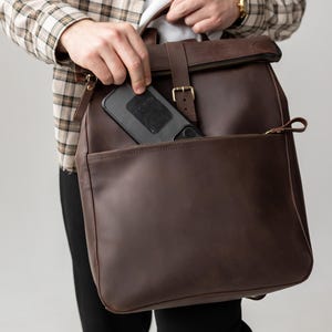 Peut inclure: Un sac à dos en cuir marron avec une fermeture à enroulement et une poche avant zippée. Le sac est doté d'une fermeture à boucle et d'une sangle. Un smartphone noir est visible à l'intérieur du haut ouvert du sac.