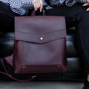 May include: A deep burgundy leather backpack featuring a structured flap with a centered brass snap closure. The front panel includes a rectangular pocket detail with two brass rivets at the bottom corners and adjustable shoulder straps.