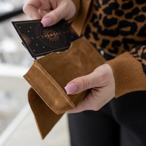 May include: A brown suede card holder with a deck of cards featuring a celestial design. The card holder is rectangular with a curved flap. The cards have a black background with a sun and moon design.