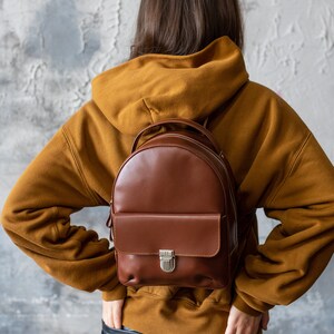 Peut inclure: Un sac &agrave; dos en cuir marron avec une poche avant et un fermoir argent&eacute;. Le sac &agrave; dos est port&eacute; sur un sweat &agrave; capuche moutarde. La personne porte un pantalon en cuir noir. Le fond est un mur textur&eacute; gris et blanc.