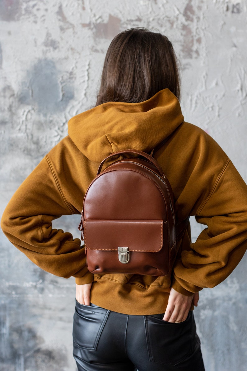 Peut inclure: Un sac &agrave; dos en cuir marron avec une poche avant et un fermoir argent&eacute;. Le sac &agrave; dos est port&eacute; sur un sweat &agrave; capuche moutarde. La personne porte un pantalon en cuir noir. Le fond est un mur textur&eacute; gris et blanc.