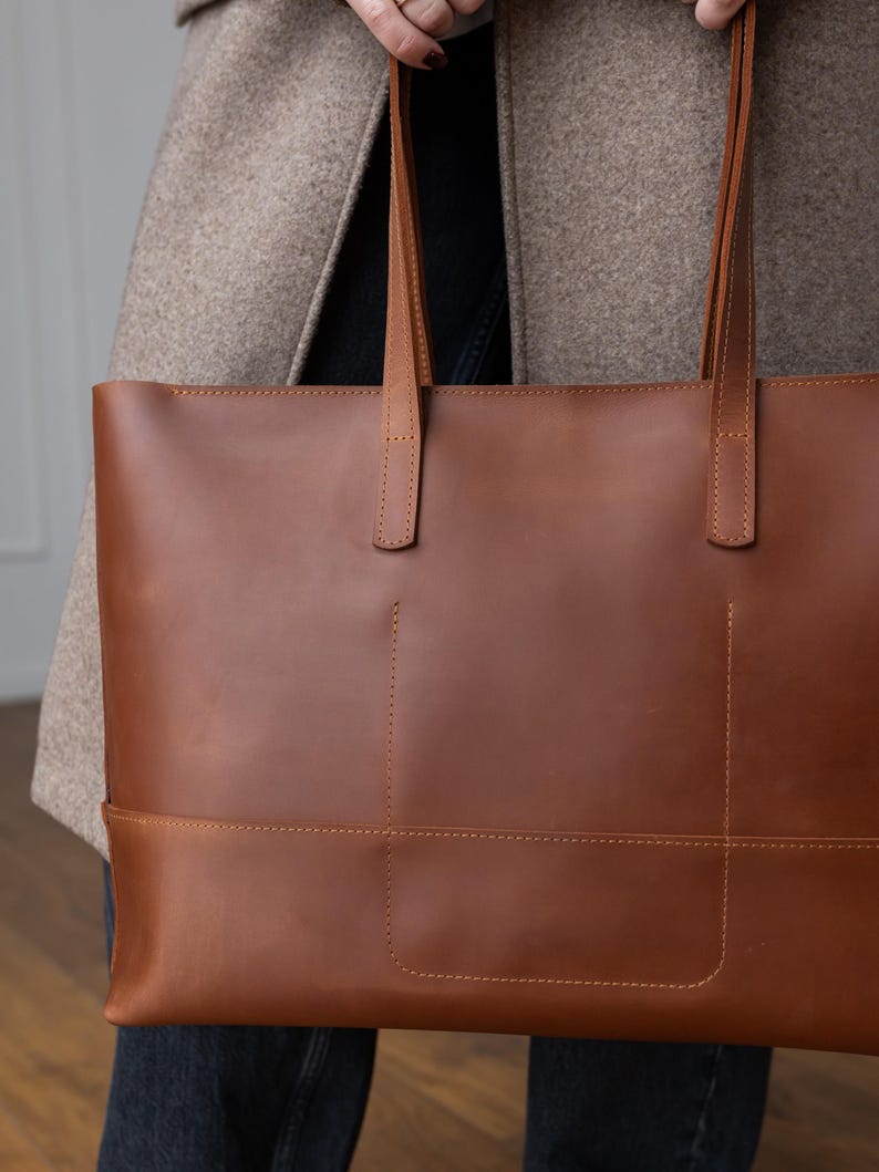 May include: A brown leather tote bag with two long handles. The bag has a rectangular shape with stitched details and a pocket on the front. The bag is held by a person wearing a tan coat.