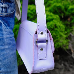 May include: A light purple shoulder bag featuring a smooth finish, a matching adjustable strap, and silver-toned metal hardware. The bag is constructed with visible stitching along the edges and is secured with metal rivets at the strap attachment points.