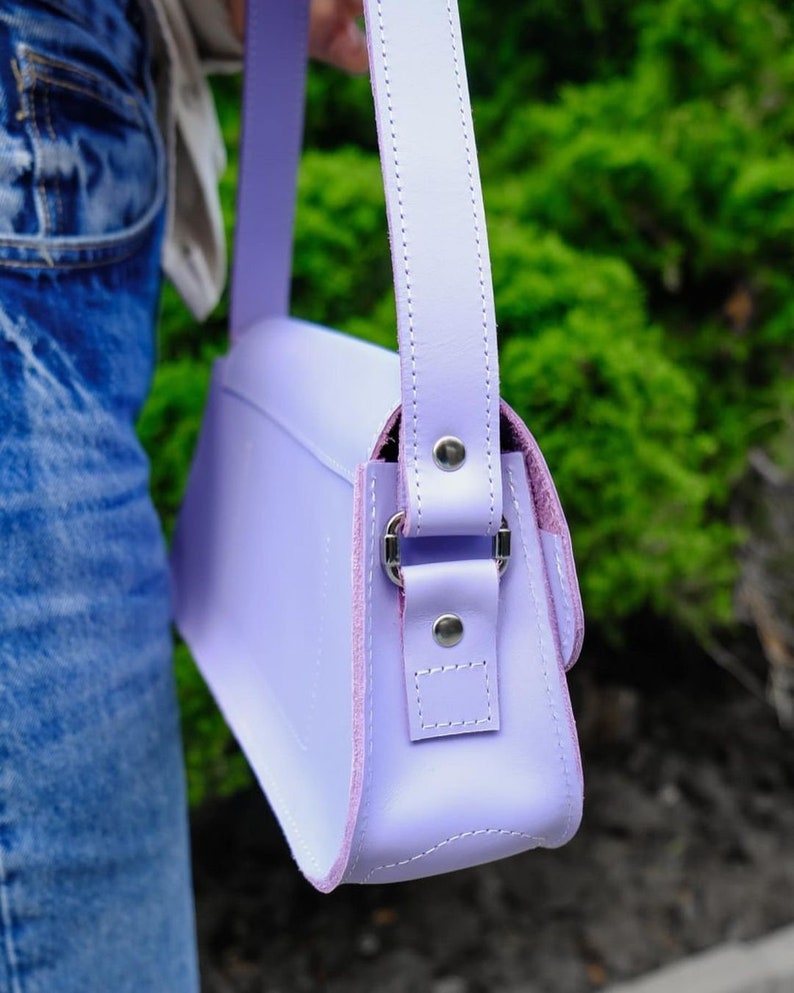 May include: A light purple shoulder bag featuring a smooth finish, a matching adjustable strap, and silver-toned metal hardware. The bag is constructed with visible stitching along the edges and is secured with metal rivets at the strap attachment points.