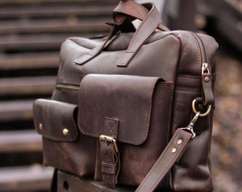 May include: A dark brown leather briefcase featuring a large main compartment with a zipper closure, two front pockets with flap covers, brass-toned metal hardware, a top handle, and a detachable shoulder strap with metal clips and rivets.