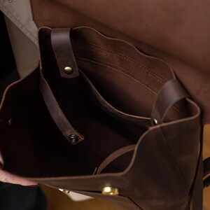 May include: A dark brown leather backpack with an open main compartment, featuring a fold-over flap, brass-toned metal snap closures, and an interior slip pocket. The bag has adjustable leather straps and visible stitching along the edges.