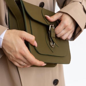May include: Olive green crossbody bag with a flap closure and a gold-colored buckle. The bag has a shoulder strap and is made of a smooth material. The bag is being held by a person wearing a tan trench coat.