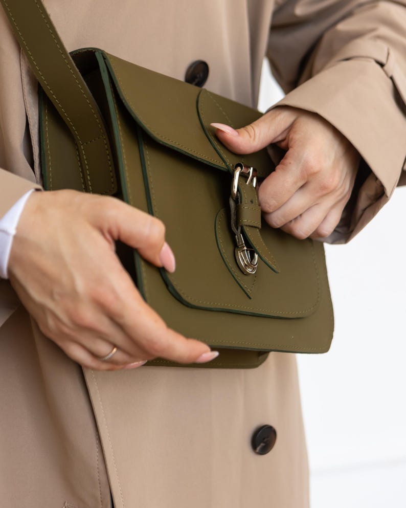 May include: Olive green crossbody bag with a flap closure and a gold-colored buckle. The bag has a shoulder strap and is made of a smooth material. The bag is being held by a person wearing a tan trench coat.