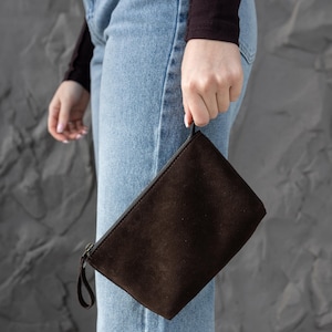 Pu&ograve; includere: Una pochette in pelle scamosciata marrone scuro con chiusura a zip e un piccolo cinturino da polso. La borsa rettangolare &egrave; tenuta su uno sfondo di jeans blu chiaro e un muro grigio strutturato.