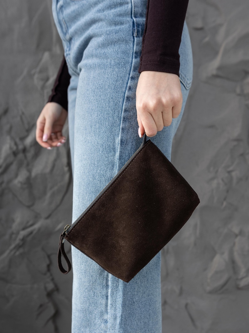 Pu&ograve; includere: Una pochette in pelle scamosciata marrone scuro con chiusura a zip e un piccolo cinturino da polso. La borsa rettangolare &egrave; tenuta su uno sfondo di jeans blu chiaro e un muro grigio strutturato.