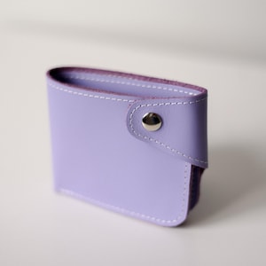 Puede incluir: Una cartera de cuero lila con costuras blancas y un cierre de botón a presión plateado. La cartera es rectangular con esquinas redondeadas y una solapa plegada. El interior es de un tono púrpura más oscuro.