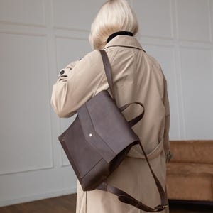 May include: A dark brown leather backpack with a fold-over flap, a single central metal snap closure, and adjustable shoulder straps. The bag features a structured rectangular shape with a top handle and is worn over a beige trench coat.