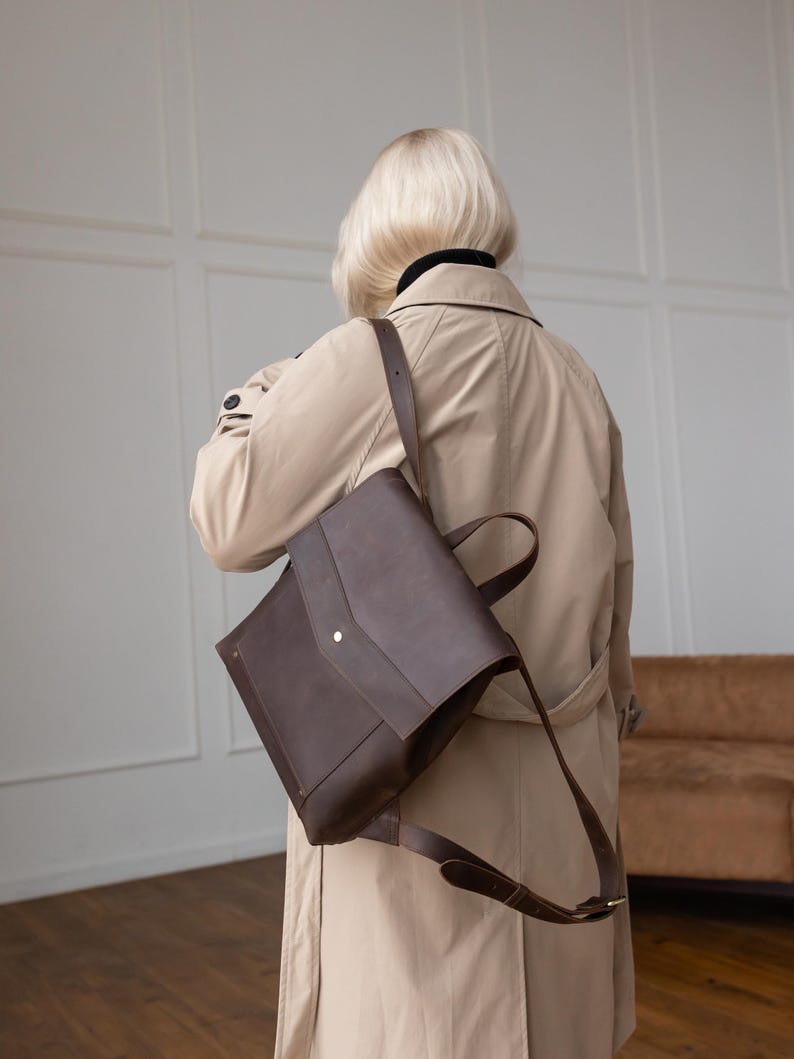 May include: A dark brown leather backpack with a fold-over flap, a single central metal snap closure, and adjustable shoulder straps. The bag features a structured rectangular shape with a top handle and is worn over a beige trench coat.