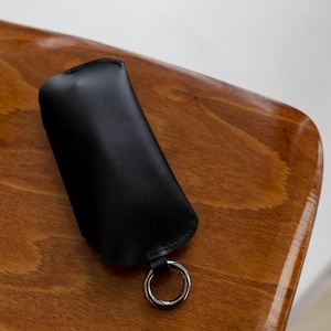 Pode incluir: Um porta-chaves de couro preto com um anel de metal prateado. O porta-chaves est&aacute; sobre uma superf&iacute;cie de madeira com um acabamento brilhante. O porta-chaves tem um design simples e minimalista.