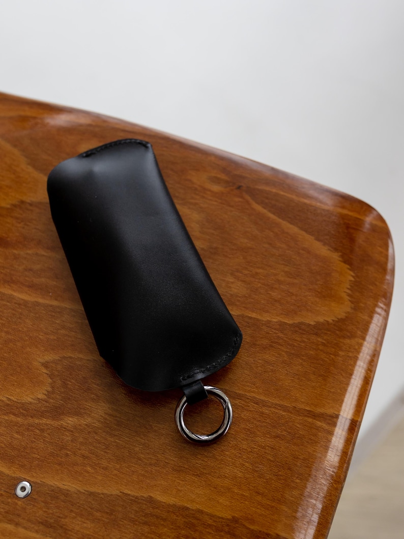 Pode incluir: Um porta-chaves de couro preto com um anel de metal prateado. O porta-chaves est&aacute; sobre uma superf&iacute;cie de madeira com um acabamento brilhante. O porta-chaves tem um design simples e minimalista.