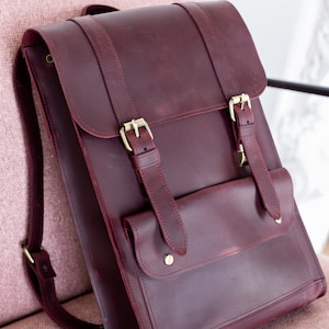 May include: A burgundy leather backpack with brass buckles and straps. The bag has a front pocket and adjustable shoulder straps. The backpack is sitting on a pink chair. The bag is a classic style.