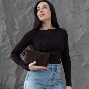 Pu&ograve; includere: Una pochette in camoscio marrone scuro con chiusura a zip. La pochette &egrave; rettangolare e sembra vuota. Lo sfondo &egrave; un muro grigio strutturato.