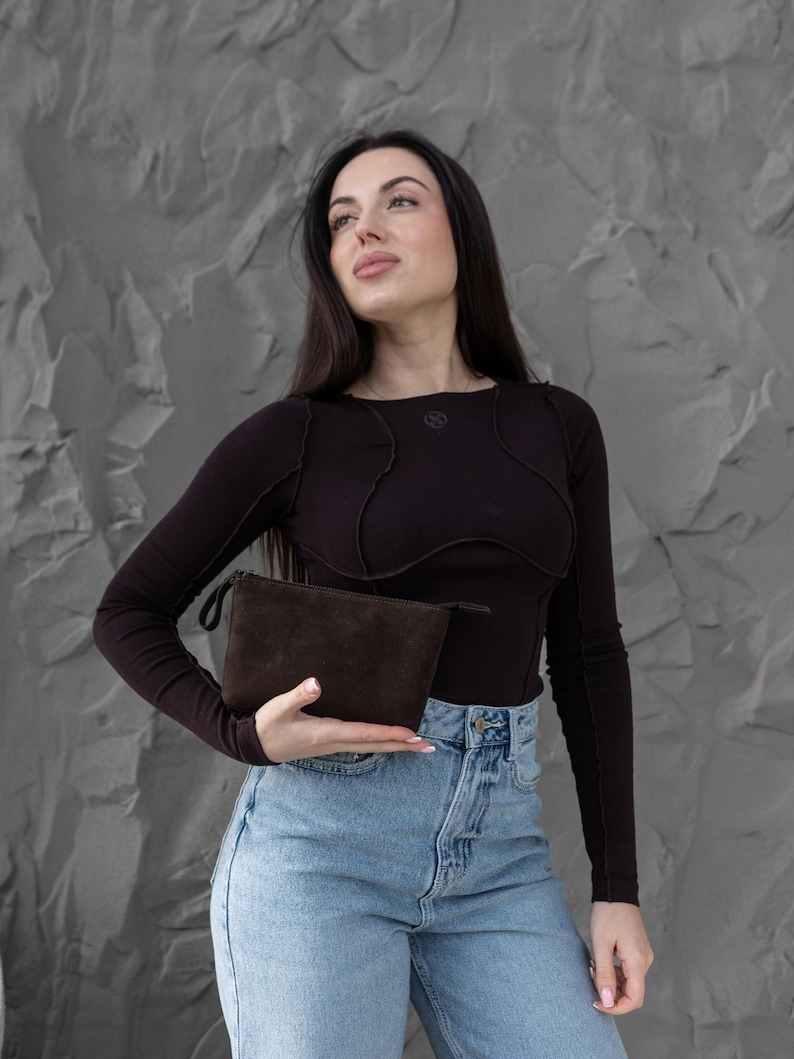 Pu&ograve; includere: Una pochette in camoscio marrone scuro con chiusura a zip. La pochette &egrave; rettangolare e sembra vuota. Lo sfondo &egrave; un muro grigio strutturato.