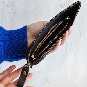 May include: Black zippered pouch with a leather pull tab. The pouch is open, revealing eyeglasses inside. The zipper and hardware are a brass color. The background is a white wall.