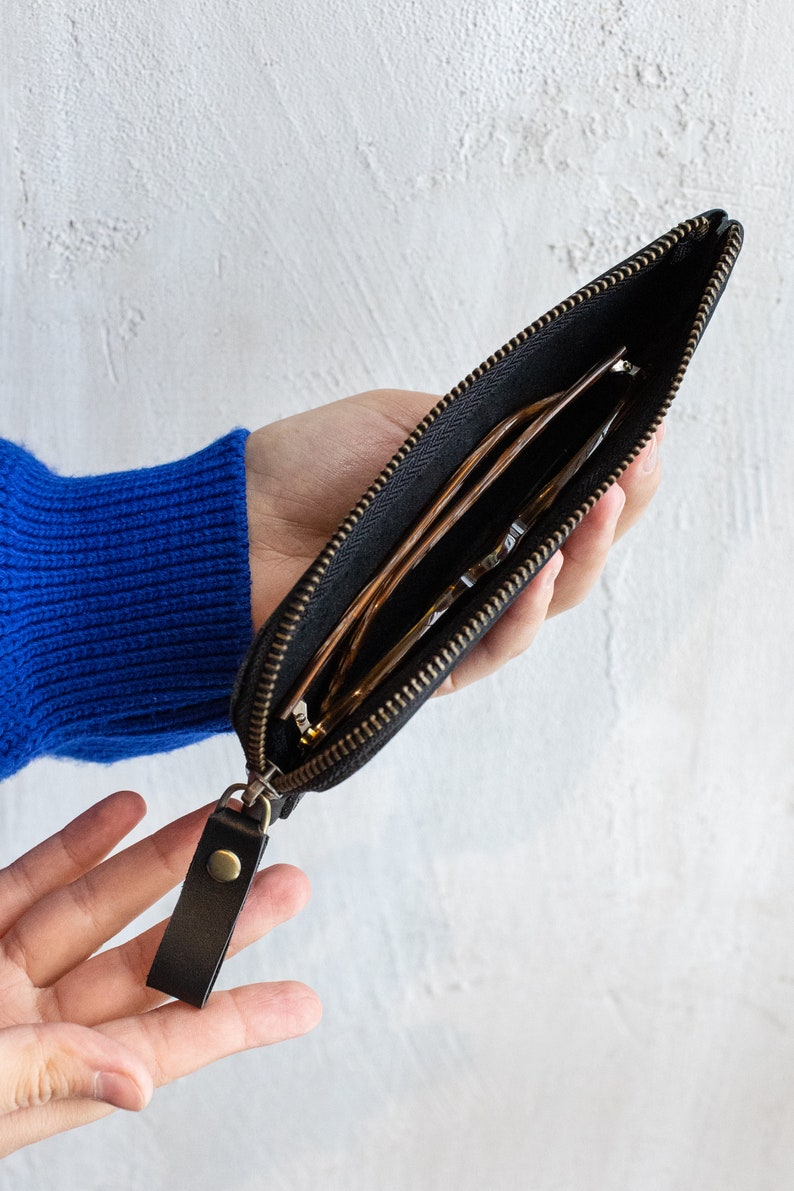 May include: Black zippered pouch with a leather pull tab. The pouch is open, revealing eyeglasses inside. The zipper and hardware are a brass color. The background is a white wall.