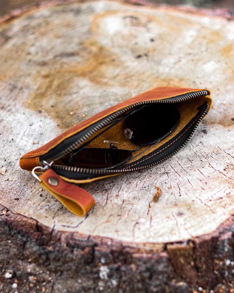 May include: A brown leather sunglasses case with a black zipper and a small leather pull tab. The case is open, revealing a pair of black sunglasses. The case is resting on a wooden surface.