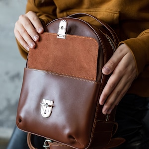 Peut inclure: Un sac &agrave; dos en cuir marron avec une fermeture &agrave; rabat et une poche avant. Le sac &agrave; dos a une poche int&eacute;rieure en daim et une quincaillerie argent&eacute;e. Le sac est tenu par une personne portant un pull moutarde.